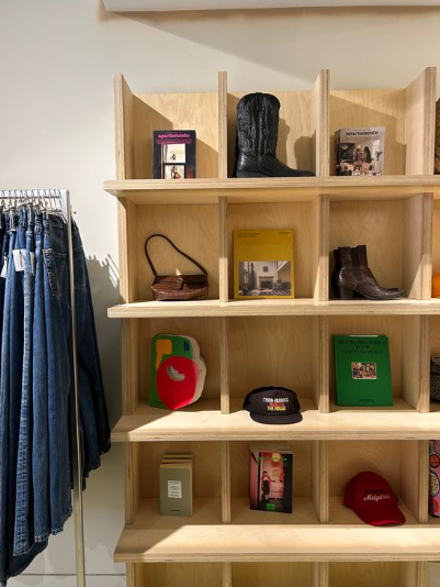 Wooden display shelves holding books, hats, bags, and boots next to a rack of blue jeans in a boutique shop.