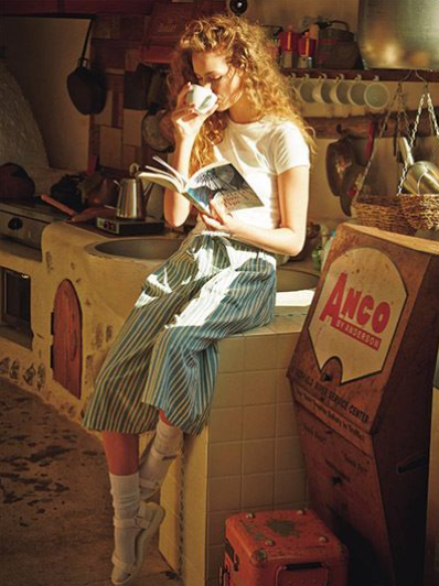A woman in a cozy, vintage-style kitchen sipping from a cup while reading a book, dressed in a striped skirt, white top, and socks.