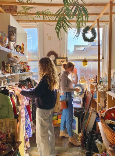 Two women browsing clothes and vintage items in a cozy, sunlit thrift shop filled with eclectic decor and racks of clothing.