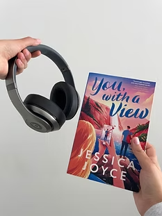 A hand holding headphones near a book titled You, With a View by Jessica Joyce.