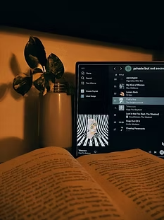 An open book with headphones placed on top, next to a laptop displaying Spotify on screen.