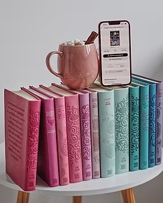 A colorful set of pink, purple, and teal hardcover books on a table, with a smartphone showing an audiobook app and a pink mug on top.