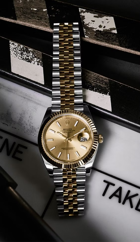A luxury Rolex watch with a gold and silver two-tone bracelet, placed on a film clapperboard. The watch face is gold with black numerals and hands, featuring a date window at the 3 o'clock position. The clapperboard has a black and white checkered pattern with the word "TAKE" visible.