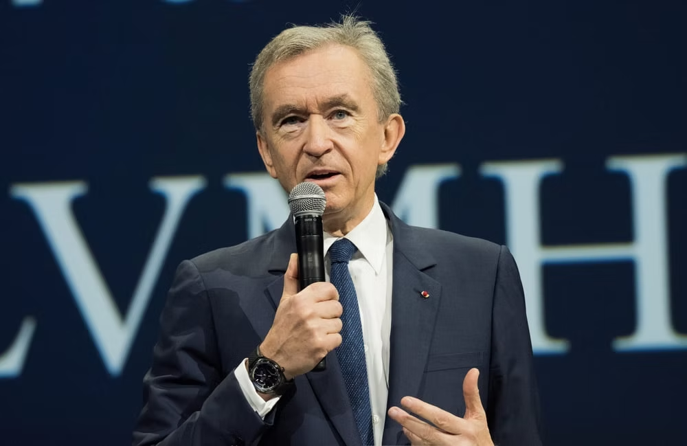 A person in a dark suit and blue tie speaking into a microphone, with a black wristwatch visible. The background features a dark blue backdrop with large white letters, partially visible.