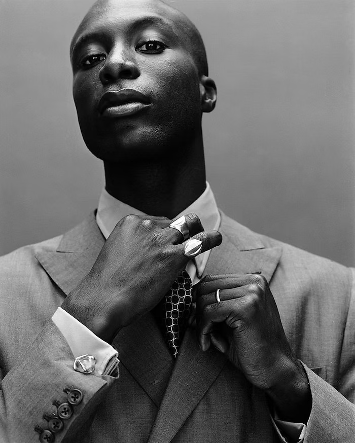 Man in a suit adjusting tie, wearing large rings. Black and white portrait with confident expression. Simple background.