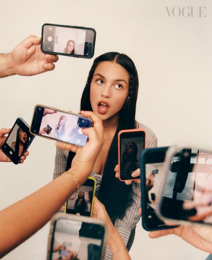 posing with an expressive face while multiple hands hold smartphones around her, all capturing photos or videos. The image, marked with the Vogue logo, conveys a theme of social media attention and digital culture.