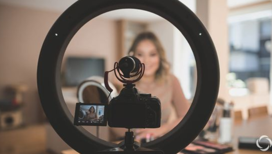 Camera setup with a ring light recording a woman in a home setting. She is slightly blurred in the background, while the camera screen shows her clearly framed for filming, suggesting a vlog or content creation session.