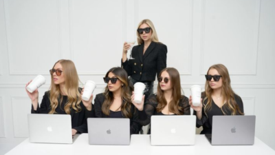 Group of women in sunglasses holding coffee cups while working on laptops, symbolizing digital culture and online content creation.