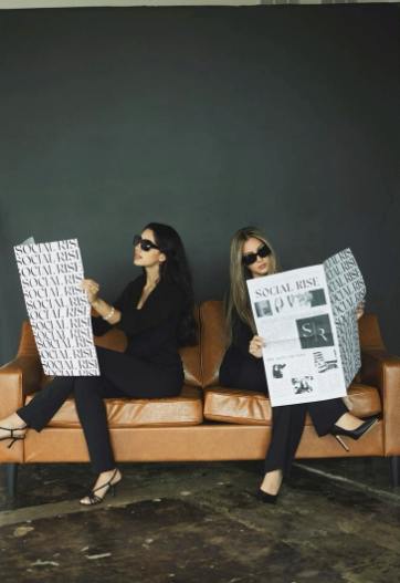 Two women dressed in black outfits and sunglasses sitting on a brown leather couch, holding large newspapers with the headline “SOCIAL RISE.”