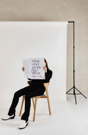 A person in black clothing and white shoes sitting on a wooden chair against a white backdrop, holding a newspaper with the headline “What other people say about us.”