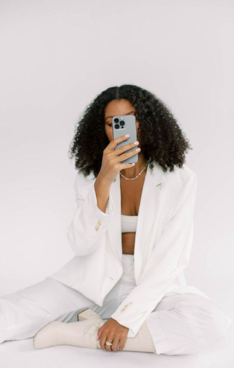 A woman in an all-white outfit sitting on the floor, holding a smartphone in front of her face as if taking a selfie or hiding her identity.