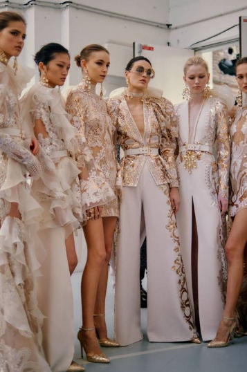 A group of models dressed in intricately designed white and gold couture outfits, standing together backstage before a runway show.