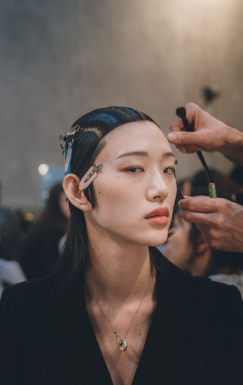 Close-up of a model getting makeup applied, her hair held in place with clips, as part of backstage preparations.