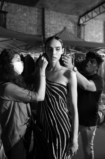 Black-and-white image of a model having final touches done on her outfit by two stylists, capturing a candid backstage moment.