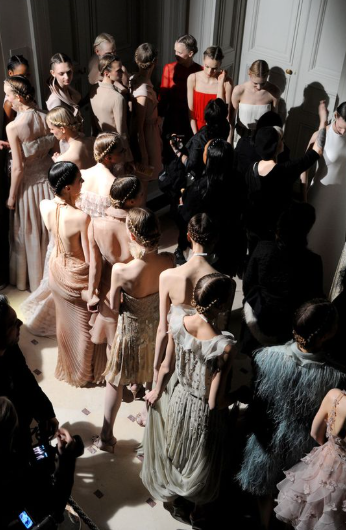 Overhead view of numerous models lined up backstage in elegant dresses, with stylists and photographers surrounding them.