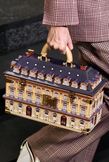 Handbag designed to look like a miniature architectural building, carried by a person in a plaid suit, symbolizing luxury craftsmanship and exclusivity in fashion.