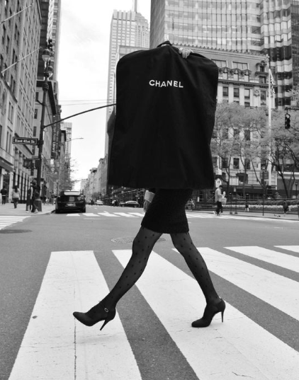 Black-and-white photo of a person crossing a city street carrying a large Chanel garment bag, with only their legs visible.
