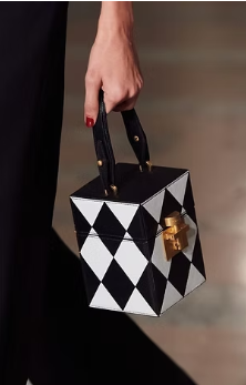 Small cube-shaped handbag with a black and white harlequin diamond pattern and a gold clasp, carried by a person in black attire.