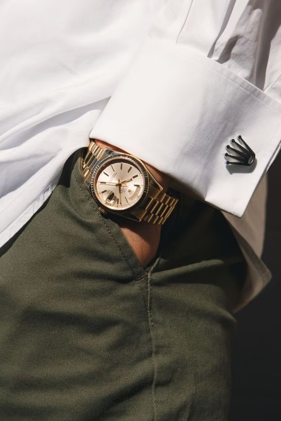 A close-up image of a person's wrist wearing a gold Rolex watch with a matching gold bracelet. The person is dressed in a white dress shirt with a black crown-shaped cufflink and olive-green trousers. The watch face is white with black numerals and hands, and a small date window is visible.