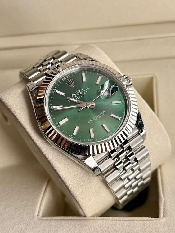A close-up image of a Rolex Datejust watch with a green dial and stainless steel bracelet, displayed on a cushioned surface inside an open box. The watch features the iconic Rolex crown logo and a date window at the 3 o'clock position with a magnifying Cyclops lens.