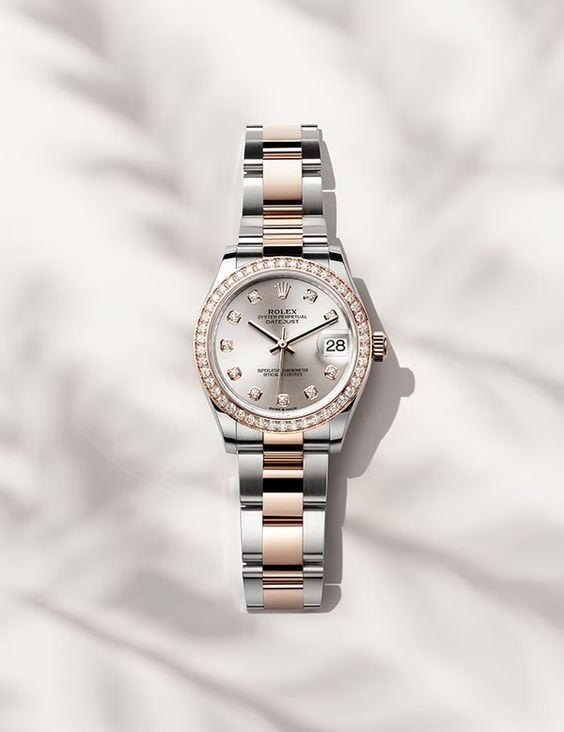 A Rolex Datejust watch with a silver dial, diamond-encrusted bezel, and a two-tone stainless steel and rose gold bracelet, displayed against a soft, light background with subtle shadow patterns. The watch features a date window at the 3 o'clock position with a magnifying Cyclops lens.