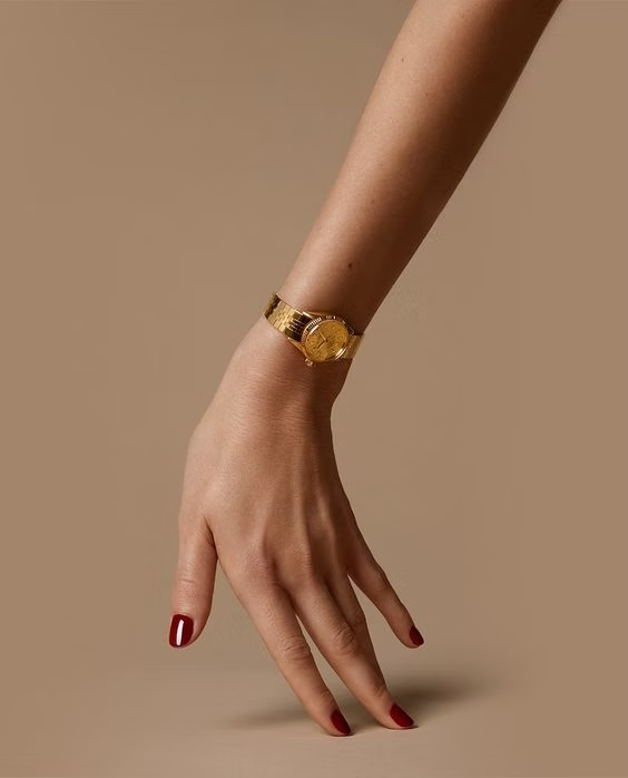 A close-up image of a person's wrist wearing a gold Rolex watch with a textured bracelet and a gold dial. The person has red nail polish on their fingernails, and the watch is set against a plain beige background.