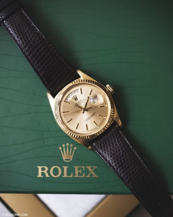 A gold Rolex Day-Date watch with a black leather strap, displayed on a green Rolex box featuring the brand's crown logo and the word "ROLEX." The watch face shows the day "FRIDAY" and the date "17" in separate windows.