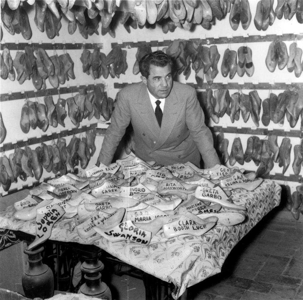 Black-and-white photograph of Salvatore Ferragamo standing in his workshop, surrounded by hundreds of wooden shoe lasts hanging on the walls. On the table in front of him are dozens of shoe lasts labeled with the names of Hollywood stars such as Greta Garbo, Gloria Swanson, and Rita Hayworth. Ferragamo, dressed in a suit, looks intently at his collection, symbolizing his legacy as the “shoemaker to the stars.”