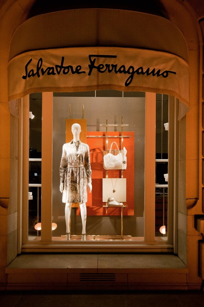 A nighttime view of a Salvatore Ferragamo store window display. A mannequin dressed in a belted, printed shirt-dress is centered against a backdrop of warm orange and neutral panels. The display also features a structured white handbag, a pair of white shoes, and a necklace, all arranged with clean geometric balance. The awning above bears the Ferragamo logo in bold black script.
