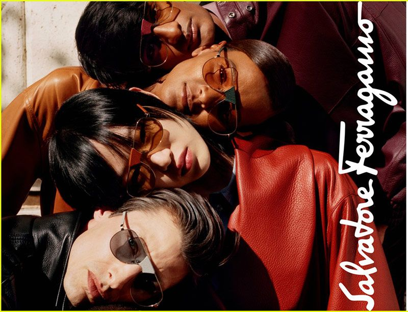 A close-up fashion campaign image featuring four models wearing Ferragamo sunglasses and leather outerwear. The models are stacked in a diagonal composition, leaning on one another, showcasing diverse skin tones and a mix of brown, red, and black leather textures. The Ferragamo logo appears vertically along the right edge of the frame.