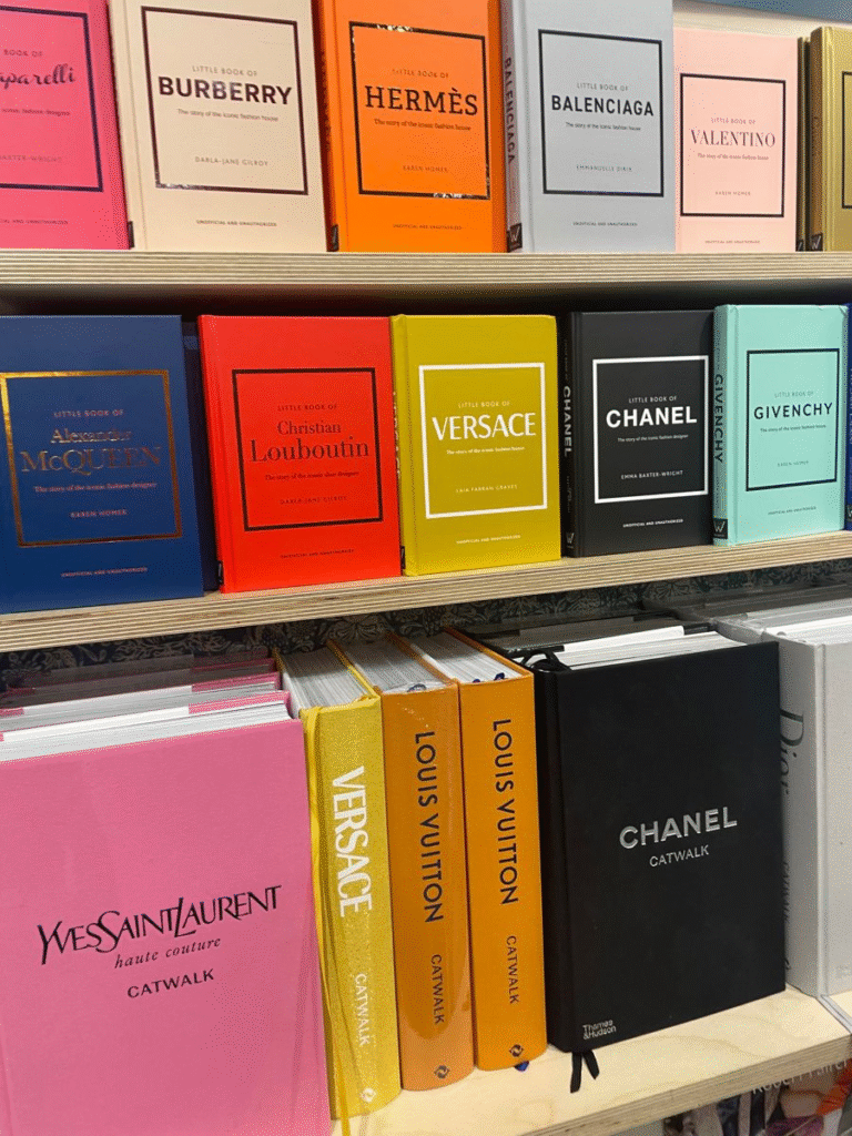 Bookshelf display featuring colorful hardcover editions of fashion books. Titles include the Little Book series on designers and brands such as Schiaparelli, Burberry, Hermès, Balenciaga, Valentino, Alexander McQueen, Christian Louboutin, Versace, Chanel, and Givenchy. Larger Catwalk series books are also visible, including Yves Saint Laurent Haute Couture Catwalk, Versace Catwalk, Louis Vuitton Catwalk, and Chanel Catwalk.