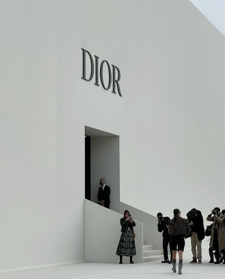 A minimalist exterior view of a Dior fashion show venue. The stark white façade is marked only by the large, elegant “DIOR” logo above a single entrance. A suited security guard stands by the doorway, while photographers, guests, and attendees gather outside. A woman in a long plaid skirt takes a photo, and another guest dressed in dark clothing walks towards the entrance.
