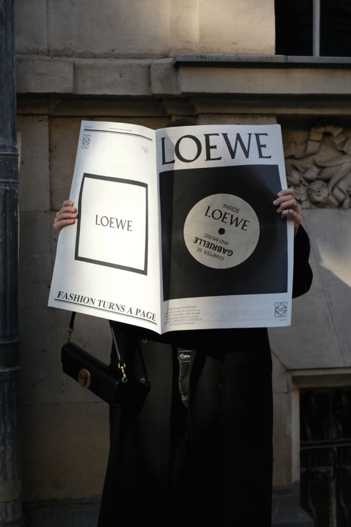 A person dressed in black holds an oversized Loewe newspaper-style publication in front of their face, obscuring their identity. The paper reads “LOEWE” in bold type, with sections including “Fashion Turns a Page” and “Inside Loewe.” The person carries a black Loewe handbag with a gold clasp, standing against a stone building wall in natural light.