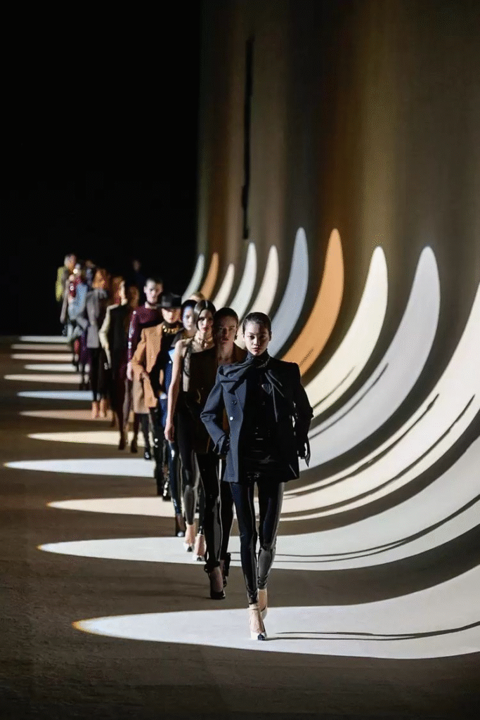 A dramatic fashion runway photograph showing a line of models walking in sequence, dressed in structured, modern outfits in a palette of black, neutrals, and metallics. The runway is illuminated by sweeping arcs of light and shadow projected onto the floor and wall, creating a striking visual rhythm that mirrors the models’ movement. The lead model wears a sharp black blazer layered over a turtleneck and skirt with high heels.