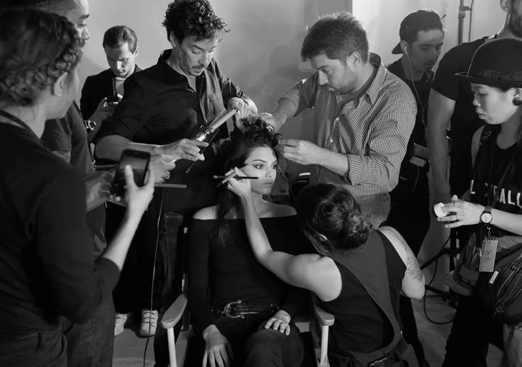 A black-and-white photograph capturing a behind-the-scenes moment at a fashion show. A model sits in a chair while multiple stylists and makeup artists work on her simultaneously — one curling her hair, another styling it, one applying eye makeup, and others assisting with details. The room is crowded with professionals focused on perfecting the look before the runway.