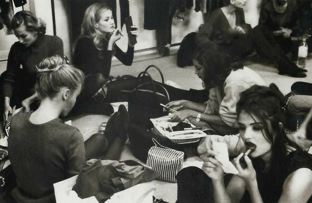 A black-and-white photograph of several models sitting on the floor backstage before a fashion show. Some are eating snacks, one applies lipstick, and others are chatting or resting with their belongings scattered around. Clothes hang on racks in the background, capturing a candid and unpolished moment behind the glamour of the runway.