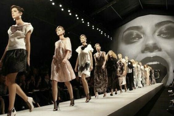 A line of models walks down the runway in succession during a fashion show finale. Each wears a different outfit ranging from light pastel dresses to dark, patterned evening looks. Behind them, a massive projected black-and-white image of a woman’s face with bold makeup and an open mouth dominates the backdrop, creating a dramatic and theatrical effect. The audience is seated in darkness along both sides of the runway.