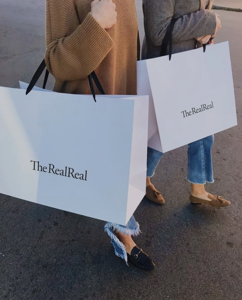 Two people carry oversized white shopping bags from The RealReal, a luxury resale company, while dressed casually in sweaters and jeans.