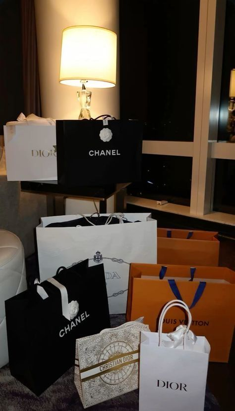 A collection of shopping bags from Chanel, Dior, Louis Vuitton, and Prada displayed in a stylish interior, with a lamp glowing softly in the background.