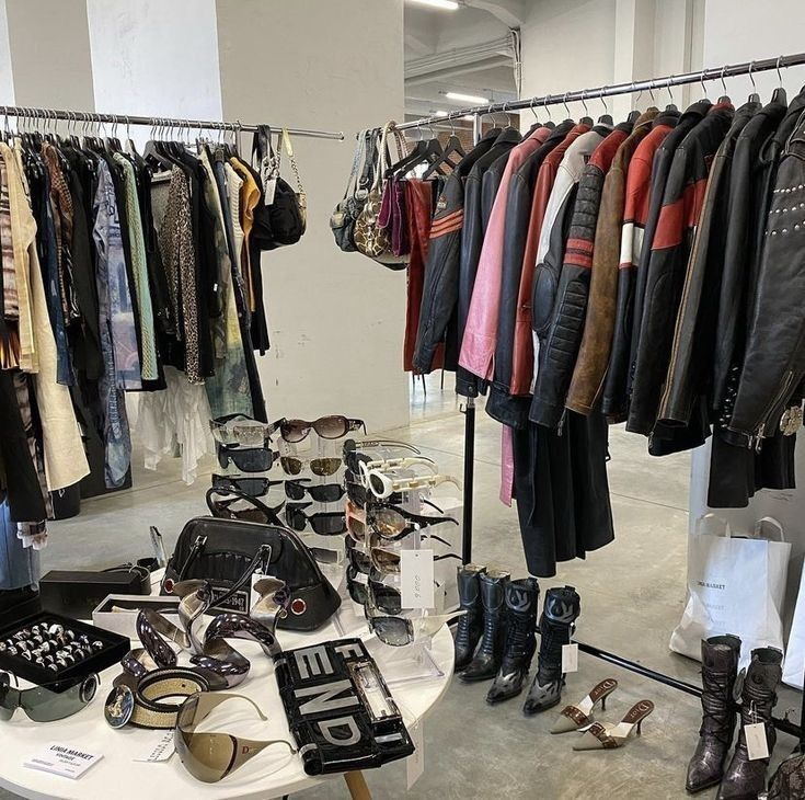 Two clothing racks filled with assorted garments and leather jackets, with sunglasses and accessories displayed in front — resembling a vintage or resale boutique setting.