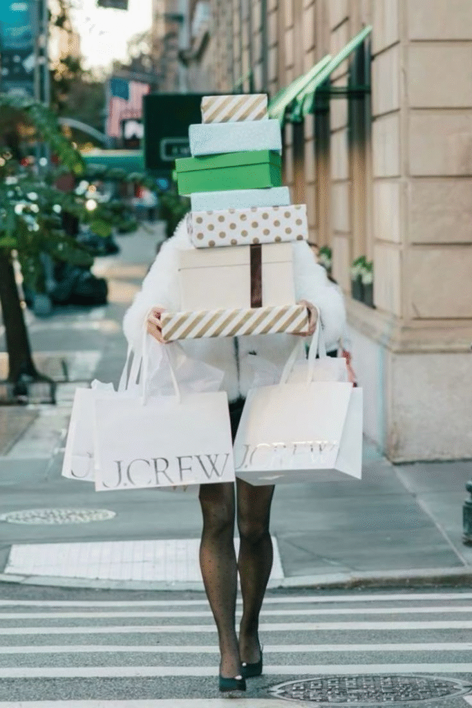 A person crosses a city street while carrying a large stack of gift boxes and multiple J.Crew shopping bags, their face obscured by the towering packages.