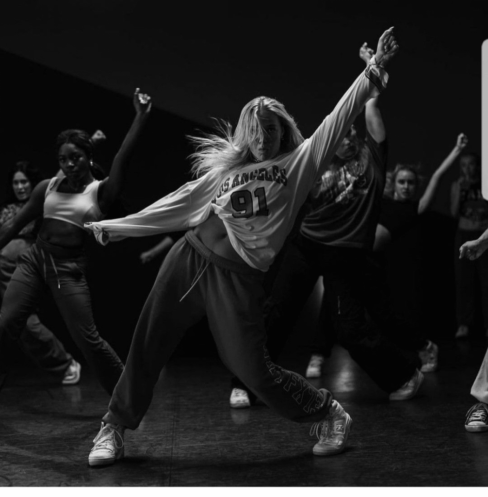 A black-and-white photo of a group of dancers in sweatpants and sneakers, mid-routine. The central dancer, in a “Los Angeles 91” sweatshirt, strikes a powerful pose with arms extended.