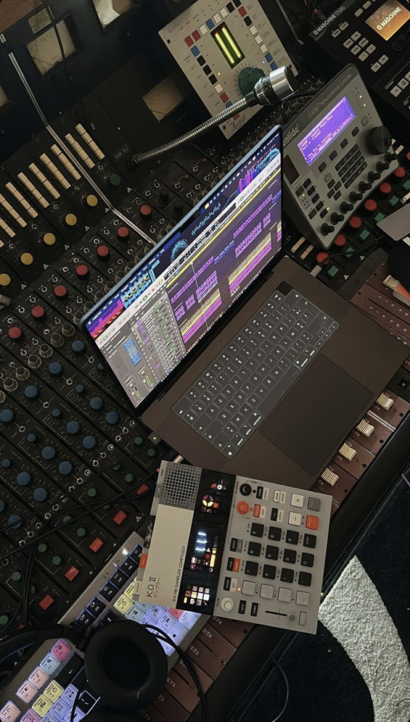 A studio desk filled with professional audio equipment, including a laptop running music production software, sound mixers, and samplers, with colorful waveforms displayed on the screen.