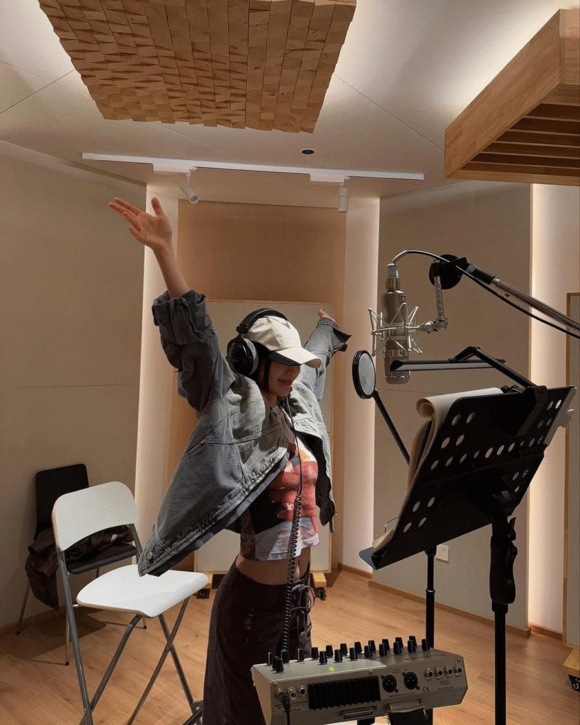 A singer in a cap, headphones, and casual streetwear stands in front of a microphone inside a recording booth, raising her arms in a celebratory gesture during a session.