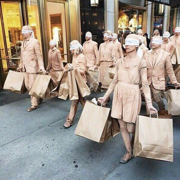 A group of people dressed in monochromatic beige clothing and blindfolds walk in unison on a city street, each carrying multiple brown shopping bags — a performance piece critiquing consumerism.