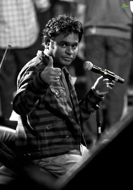 A man in a striped jacket gives a thumbs-up while holding a microphone on stage, captured in black-and-white with a spotlight effect.