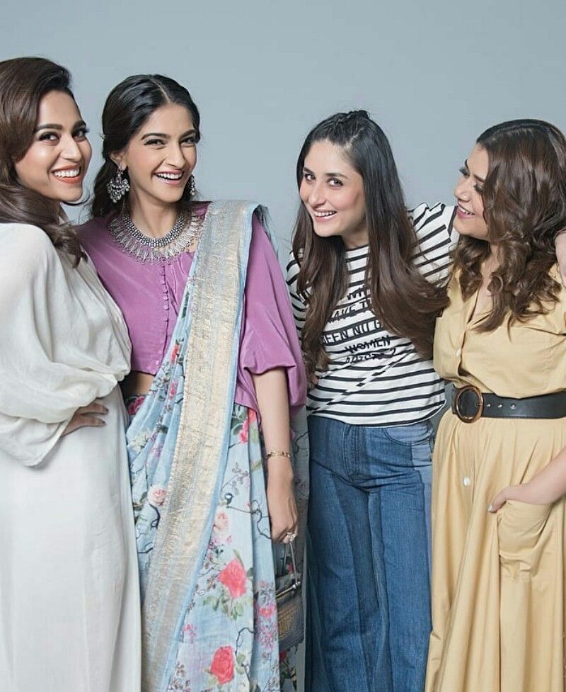 Four women standing together, smiling and laughing. The woman on the left wears a long white dress, the second woman wears a purple blouse with a floral blue saree, the third woman wears a striped black-and-white top with wide blue jeans, and the fourth woman wears a belted mustard-yellow dress. They have their arms around each other in a friendly pose against a plain grey background.