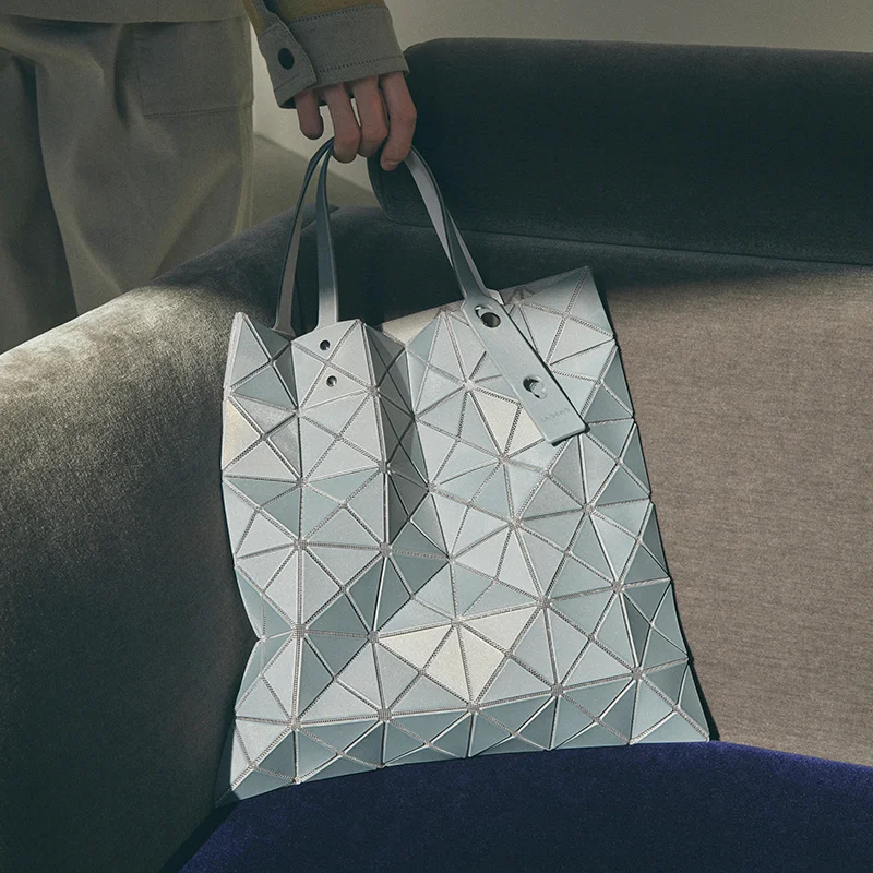 Close-up of a BAO BAO ISSEY MIYAKE tote bag resting on a sofa. The bag features a signature geometric design made of interconnected triangular panels in a light gray shade, with two structured handles.