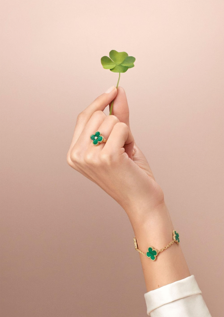 A graceful hand holds a fresh green four-leaf clover against a soft blush background. On the ring finger is Van Cleef & Arpels’ Alhambra ring in gold with malachite, while a matching bracelet featuring the same clover motif encircles the wrist — a refined tribute to the collection’s 50-year legacy of luck and timeless elegance.