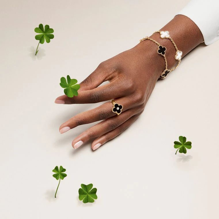 A poised hand rests on a light surface, surrounded by scattered four-leaf clovers. The fingers are adorned with Van Cleef & Arpels Alhambra jewelry — a gold ring with a black onyx clover and two matching chain bracelets featuring alternating motifs in black onyx and mother-of-pearl. The scene evokes timeless charm and the collection’s emblematic symbol of luck.